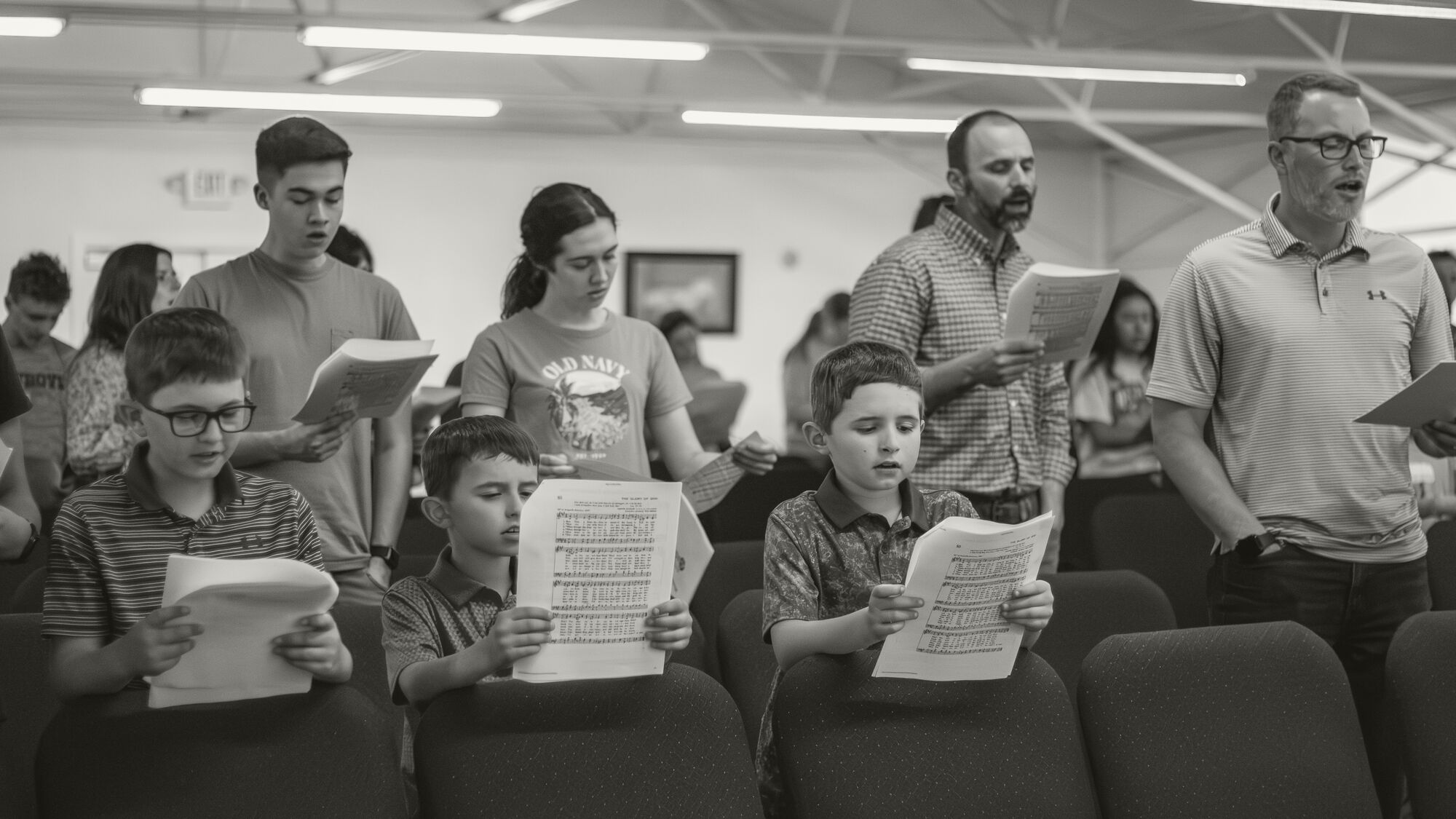 Three boys in the front row sing from sheet music while teenagers and men sing behind them.